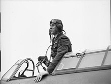 Una fotografía en blanco y negro de un hombre de pie en la cabina de un avión;  el usa un casco volador