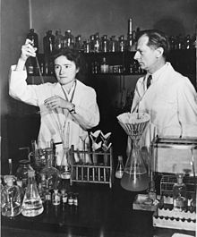 Fotografia em preto e branco que mostra um homem e uma mulher com jalecos em um laboratório.