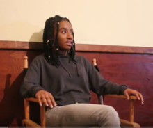 Hakim sits in a chair against the wall. She has brown skin, her hair is in twists, and she is wearing a hoodie.