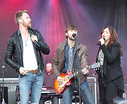Two men and a woman performing on a stage, both singing into microphones. All are holding microphones and one of the men is also playing a guitar.