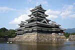 A large 5-storied castle tower with black wooden walls located on a platform of unhewn stones surrounded on two sides by water. The tower is connected to lower structure on two sides.