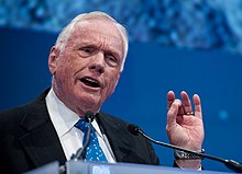 An elderly but fit-looking Armstrong in mid-speech. He is wearing a dark suit, a white shirt and a pale blue tie. He holds up his left hand and touches the thumb to the middle finger.