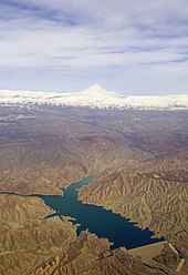 Vue aérienne un lac de retenue au bleu profond, des montagnes ocres dénuées de végétation, en arrière-plan des crêtes plus hautes et un cône volcaniques enneigés.
