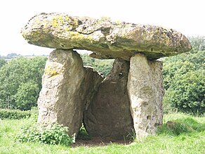 Front view of a dolmen. Its massive capstone is supported by standing stones to either side, with another (triangular) supporting stone at the rear – like a doorless closet. The rear orthostat has a small round hole near the middle top. The dolmen is set in an open, sloping (higher&ndashleft, lower–right) meadow of uncut grass, with trees to the rear in the middle distance.