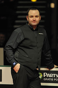Zdjęcie. Mężczyzna ubrany w czarny garnitur do gry w snookera spogląda w kierunku fotografującego.
