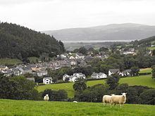 Un petit village au milieu de vertes collines arborées où paissent des moutons au premier plan.
