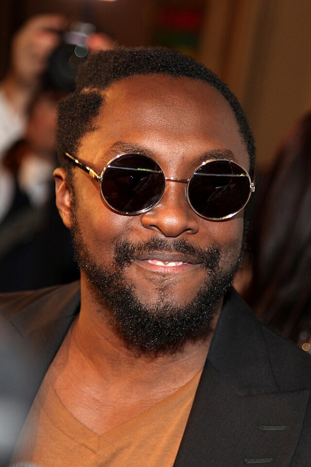 A dark-skinned man with black hair and black facial hair is seen wearing glasses with circular lenses.