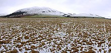 Fotografía de un campo en barbecho cubierto de nieve, con picos montañosos en el horizonte.