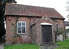 Close view of a simple single-storey building with two windows, a porch, alternating red and grey bricks and some gravestones below the windows.