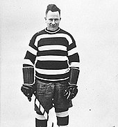 Black and white photo of a hockey player wearing hockey equipment, holding a stick, and wearing a horizontal-striped jersey