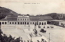 Schwarzweiss-Postkarte eines horizontalen Gebäudes mit einem großen Vorplatz, vor dem Wagen sind.