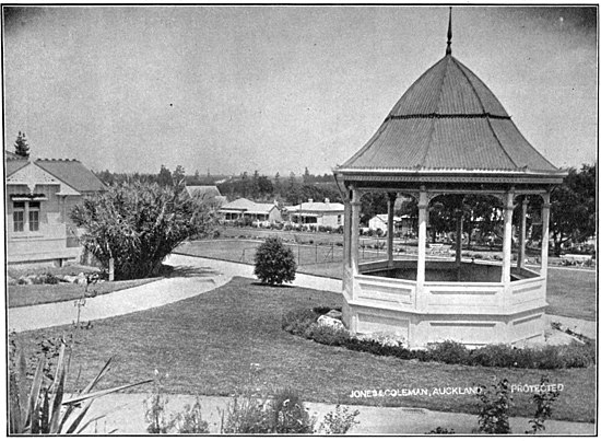 A bandstand in the front-right foreground, lawn with path and a house to the left, and other houses and trees in the background