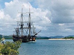 A three-masted wooden sailing ship crossing a calm bay, beneath a cloudy sky.