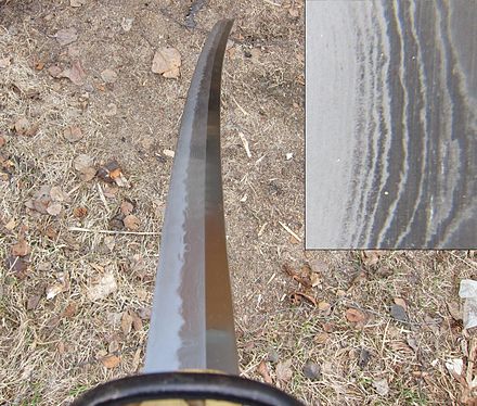 A katana, shown at a long angle to reveal the nioi, which is the bright, wavy line following the hamon. The inset shows a close-up of the nioi, appearing as the speckled area beside the bright hardened-edge. The nioi is made up of niye, which are single martensite crystals surrounded by darker pearlite.