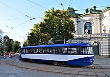 Seitliche Farbfotografie einer blau-weißen Straßenbahn, die über eine Kurve in eine Allee mit alten Gebäuden fährt. Rechts hinten ist ein weißes Gebäude mit rundem Mittelteil und grünem Dach sowie Ornamenten.
