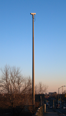 "A video camera mounted on a tall cement pole which is mounted on the side of a roadway. The camera is not pointing at the roadway visible at the bottom-right of the picture, but to the left."