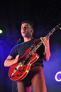 Млад мъж от Кавказка, който държи червена електрическа китара, застава на тъмен фон, осветен със син прожектор.