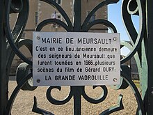 Photographie en plan rapproché d'un portail en fer forgé sur lequel est apposé une plaque blanche indiquant « MAIRIE DE MEURSAULT - C'est en ce lieu, ancienne demeure des seigneurs de Meursault, que furent tournées en 1966, plusieurs scènes du film de Gérard OURY : LA GRANDE VADROUILLE ».