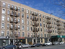 An apartment building complex with multiple square windows that are right beside fire escapes. In front of the building are six cars, of different colors, and a street.