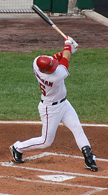 Un hombre, agarrando un bate de béisbol y con un casco de bateo rojo y un uniforme de béisbol blanco con su apellido y número parcialmente oscurecidos en su espalda, se balancea en un lanzamiento.