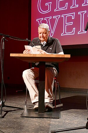 Ganzkörperbild eines Mannes mit kurzen grauen Haaren, der an einem Lesetisch sitzt und in ein Standmikrofon spricht. Er hält ein schmales weißes Buch aufgeschlagen in den Händen.