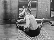 Un homme, assis jambes écartées au sol, devant un miroir, attrape la pointe de son pied gauche avec sa main droite.
