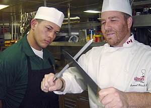 US Navy 031204-N-1711I-001 Mess Management Specialist 3rd Class Benjamin AlmodovarPacheco from Ceiba, Puerto Rico, watches carefully as culinary arts instructor Chef Sam Glass trains him on the best way to sharpen a butcher's.jpg