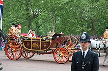 Couple sitting in a decorated horse-drawn open-top carriage, with two well-dressed guards sitting behind the newly-weds. The carriage is flanked on the far side by excited well-wishers