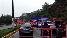 صحنه‌ای از یک خیابان نسبتاً شلوغ با تعدادی اتوموبیل سواری و موتور سیکلت در آن که هر یک توسط یک الگوریتم بینایی ماشین رهگیری شده، مستطیلی به دورش رسم شده و برچسبی به بالای مستطیل زده شده است.