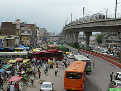 Röd och orange färgade Delhi Transport Corporation komprimerade naturgasbussar mitt i gångtrafik under det förhöjda tunnelbanetåget i Delhi