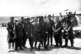 Nine men wearing a mixture of military uniforms with caps and flying suits with goggles, in front of a row of military biplanes