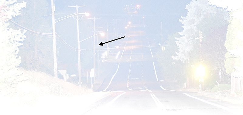This photo was shot in fair quality air at ISO 12,800 using a modestly powerful camera-mounted flash, yielding a high guide number of 438 (m) / 1438 (ft). When shot at f/1.8 to favor distance, the utility pole marked by the arrow would be properly illuminated were it not for haze glare, which fogged the image and diminished brightness.