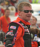 A man in his forties wearing black sunglasses and an orange-and-black jacket with sponsors logos