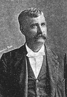 Un homme dans la quarantaine avec des cheveux noirs et une longue moustache portant une chemise blanche et un manteau noir, tourné vers la droite