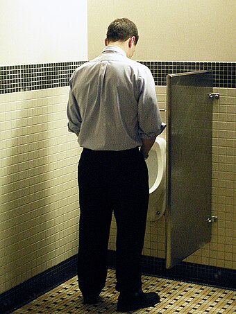 A man uses a urinal while urinating in a standing position.
