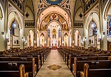 Interior view from the rear of the center aisle of a large, well lit, neo-Gothic church, toward the colorful apse.