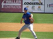 Мужчина в серых штанах и синей бейсбольной майке с надписью «METS» на груди готовится подать бейсбольный мяч правой рукой.