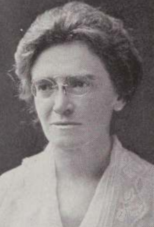 A middle-aged white woman wearing pince-nez glasses and a white blouse or dress with a lace collar