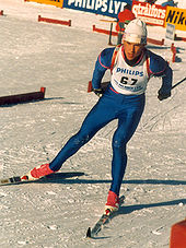 Ein Mann fährt auf einer schneebedeckten Strecke.Er trägt eine weiße Weste über einem blauen, hautengen Overall, eine weiße Wintermütze und rote Skischuhe.Auf dem Rücken trägt er ein Gewehr.