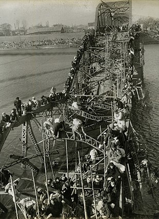 Schwarzweißfoto von Menschen, die einen Fluss über eine zerstörte Brücke überqueren