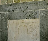Photographie d'un panneau en marbre blanc, bordé de bandeaux verts, qui se trouve à gauche de la niche du mihrab. Il est sculpté d'un arc recti-curviligne, dont l'un des écoinçons est orné d'une inscription coufique rayonnante. Celle-ci est inscrite dans un cercle orné.