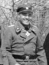 Un homme souriant vêtu d'un uniforme militaire, casquette à visière avec emblème du crâne et ordre du cou en forme de croix.