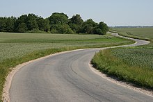 Pole lub łąka, przez które przechodzi wąska utwardzona droga. Jest kręta, nie ma na niej namalowanego oznakowania. Nie jedzie nią żaden samochód.