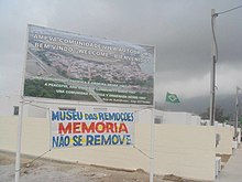 Placa na entrada da Rua Vila Autódromo. Abaixo da placa, faixa com tipografia vernacular apresenta a mensagem "Museu das Remoções: Memória não se remove".