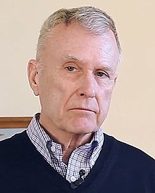 Richard Nisbett wearing a dark sweater, with a light checkered shirt visible underneath, scowling and looking right of camera