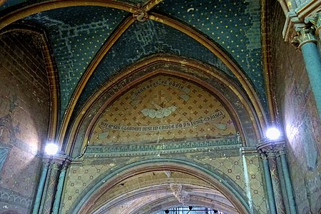 Photographie d'un tympan peint avec des inscriptions latines et deux bras croisés devant une croix et des nuages. Au-dessus, des voûtes peintes en bleu et jaunes. De part et d'autres, des piliers avec des lampes allumées.