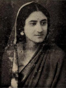 A South Asian woman with fair skin and dark hair parted center, wearing a scarf draped loosely over her head