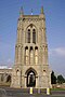 A tower with an open lower stage, and arches in the stages above and on the buttresses; at the top is a decorated parapet and crocketted pinnacles