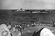 Black and white photo of two aircraft carriers sailing in close formation viewed from the flight deck of another aircraft carrier