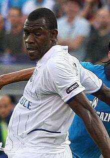 Hombre de color negro, vestido con una camiseta blanca, brazos detrás de la espalda y mirando al suelo